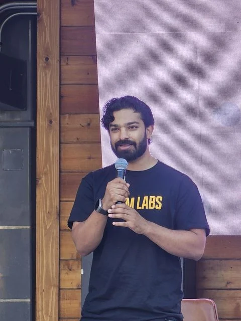 Aditya Goyal speaking into a microphone on stage, wearing a Prism Labs shirt